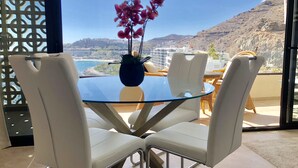A modern dining area with a glass table and white chairs, overlooking a scenic coastal view. - Wake up to Gorgeous Ocean Views from this Chic Apartment (Patalavaca)