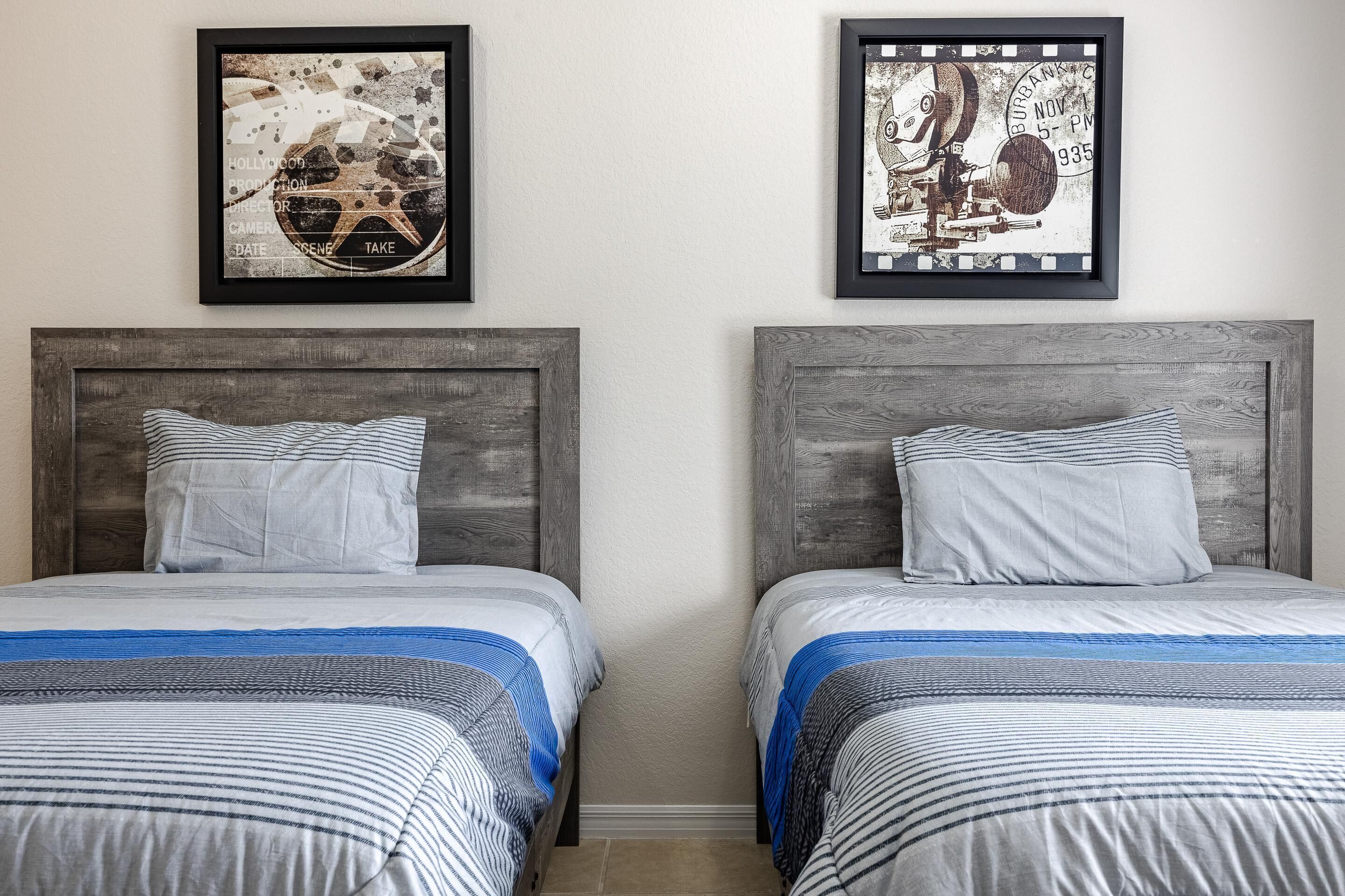 A hotel room with two beds, each with a headboard, a striped bedspread, and a pillow. There are two framed pictures on the wall above the beds.