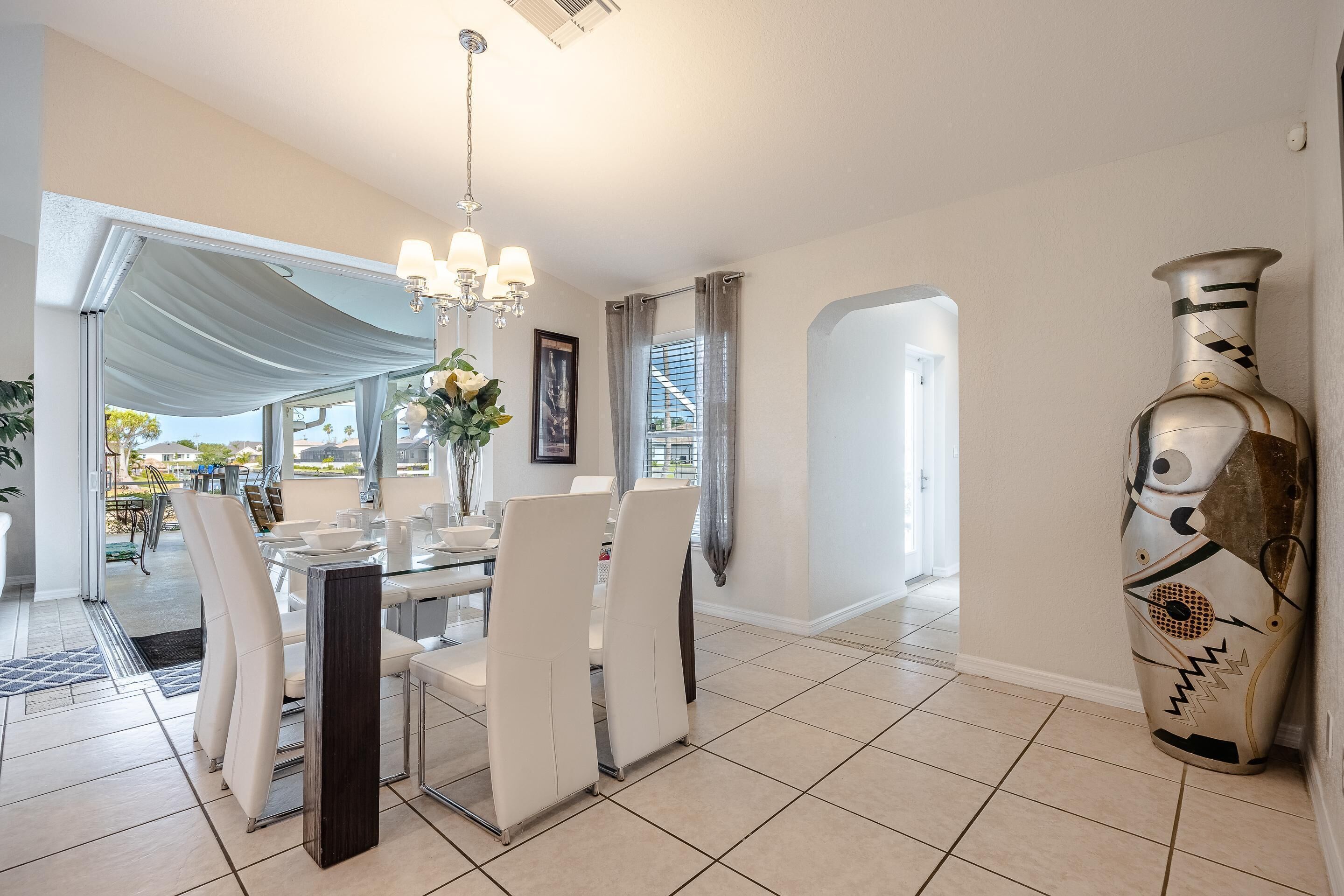 A dining area with a table set for four, white chairs, a chandelier, and a large vase on the wall.