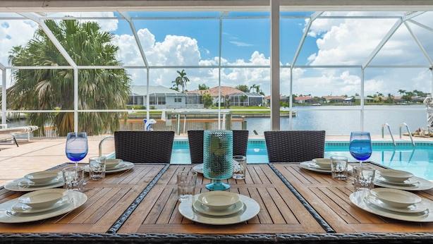 Uma área de refeições ao ar livre com vista para um lago, mesas de jantar montadas com pratos e copos, e um enfeite de vidro decorativo.