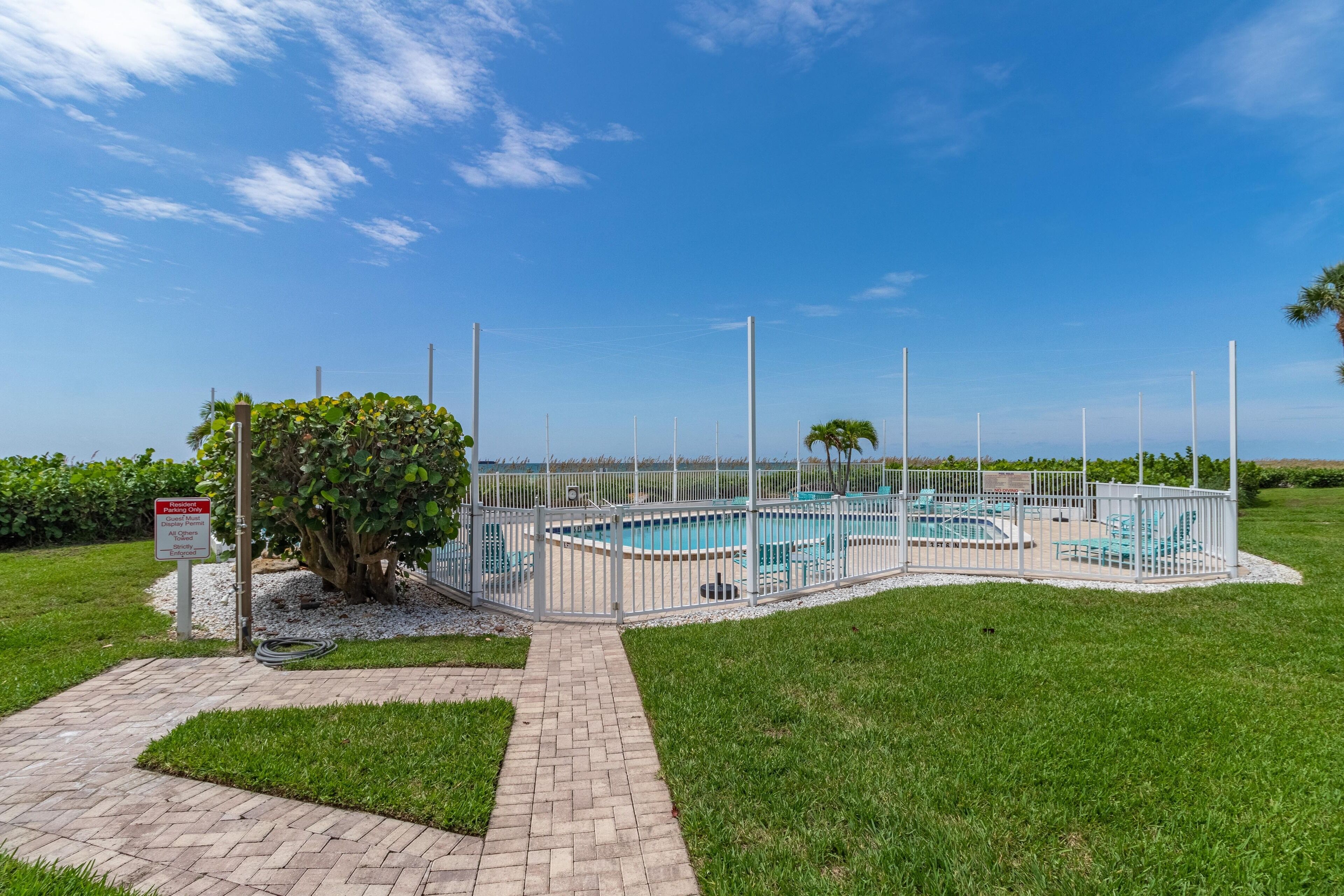 A gated swimming pool area with a paved walkway leading to it, surrounded by greenery and a clear sky.