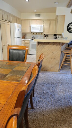 A kitchen with wooden cabinets, a white refrigerator, a microwave, and a stove. There is a dining table with chairs and a carpeted floor.