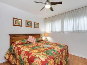 A bedroom with a floral bedspread, a ceiling fan, and three framed pictures on the wall. - Modern Hawaii Style w/Kitchen, WiFi, Lanai–#205 (KAUNAKAKAI)