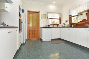 A compact kitchen with white cabinets, a wooden door, a window with lace curtains, and a tiled floor. - La Sila Homestead on Marrowbone Great Views (Pokolbin)