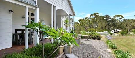 A white house with a covered porch, outdoor seating, and a garden with various plants and a gravel pathway.
