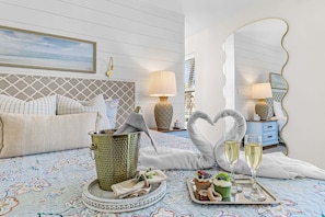 Een slaapkamer met een bed, nachtkastje, lamp, spiegel, dienblad met drankjes en gebak, en decoratieve, zwaanvormige kussens.