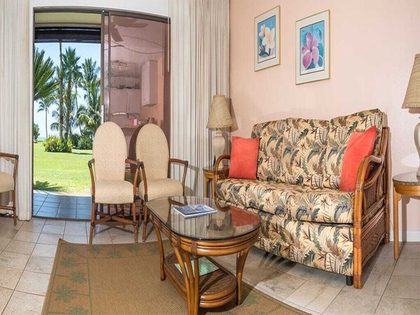 Een woonkamer met een bank, twee fauteuils, een glazen salontafel en uitzicht op palmbomen door een schuifdeur.