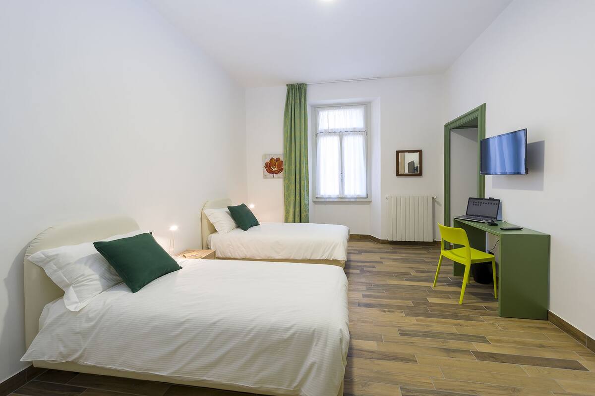 Une chambre avec deux lits, un bureau avec un ordinateur portable, une chaise, une télévision et une fenêtre avec des rideaux.