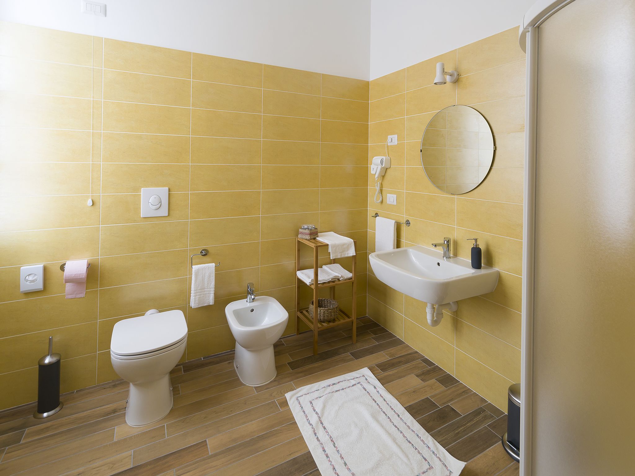Une salle de bain avec des carreaux jaunes, un lavabo blanc, un bidet, des toilettes, un miroir et une étagère en bois avec des serviettes.
