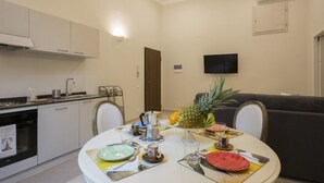 Una zona de menjador amb una taula rodona preparada per a quatre, una cuina amb armaris blancs, un televisor de pantalla plana fixat a la paret i un sofà de color fosc.
