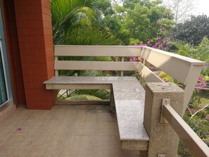Un banco de hormigón con estructura metálica, ubicado en un balcón con suelo de baldosas, un muro de ladrillo rojo y una puerta de cristal.