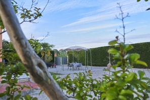 Une terrasse extérieure avec une table et des chaises abritées, entourée d’une haie et d’arbres.