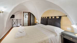 Une chambre à coucher avec un plafond en arc de cercle, un lit recouvert de draps blancs, une table de chevet et un téléviseur fixé au mur.