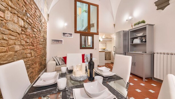 Un coin repas avec un mur en pierre, une table en verre, des chaises blanches, une bouteille de vin et, au fond, une cuisine.