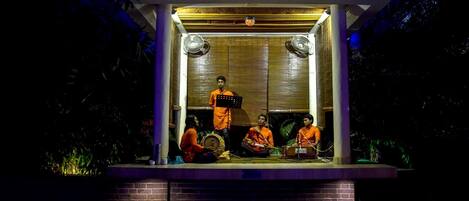 Una pequeña estructura iluminada con techo, posiblemente un pabellón, con tres personas adentro: una sentada, otra de pie con un atril musical, y un abanico en la pared.