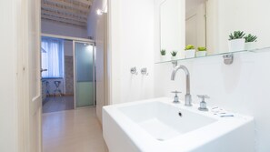 Une salle de bain moderne avec un lavabo blanc, une étagère en verre ornée de plantes et un espace douche visible à travers une porte en verre.