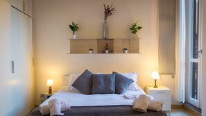 Un lit bien fait, avec des oreillers et une couverture, des lampes de chevet, une petite étagère avec des plantes et une fenêtre avec des rideaux.