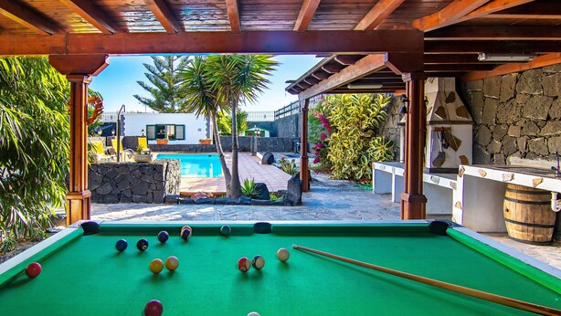 A pool table with balls, a stone wall, and a pool in the background. - Villa Leonard in Playa Blanca (Playa Blanca)
