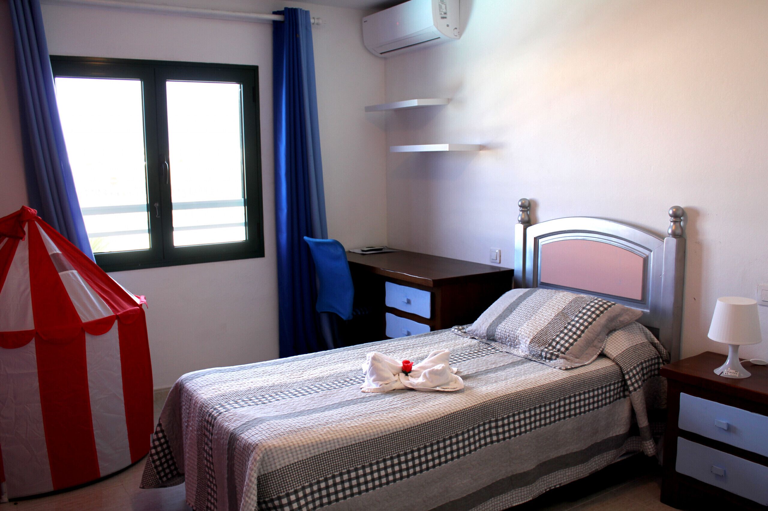 A hotel room with a bed, a desk, a chair, a wardrobe, and an air conditioning unit.