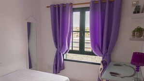 A room with a bed, a window with purple curtains, a mirror, a small table with a lamp, and a chair. - Villa Leonard in Playa Blanca (Playa Blanca)