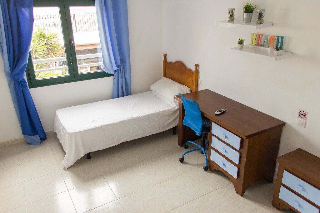 A bedroom with a single bed, a wooden desk with drawers, a chair, and a shelf with decorative items.