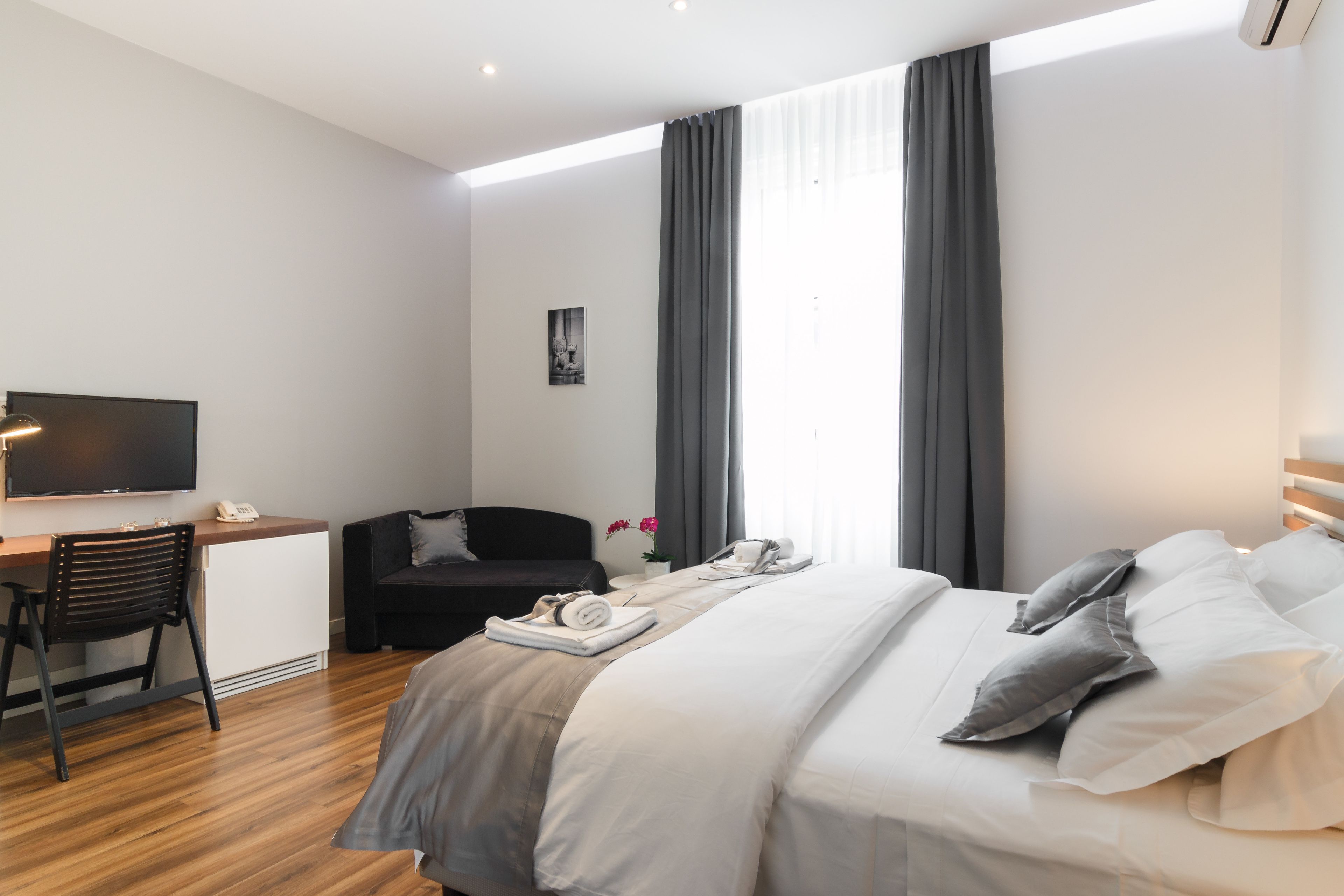 A modern hotel room with a bed, a desk, a chair, a TV, and a window with curtains.