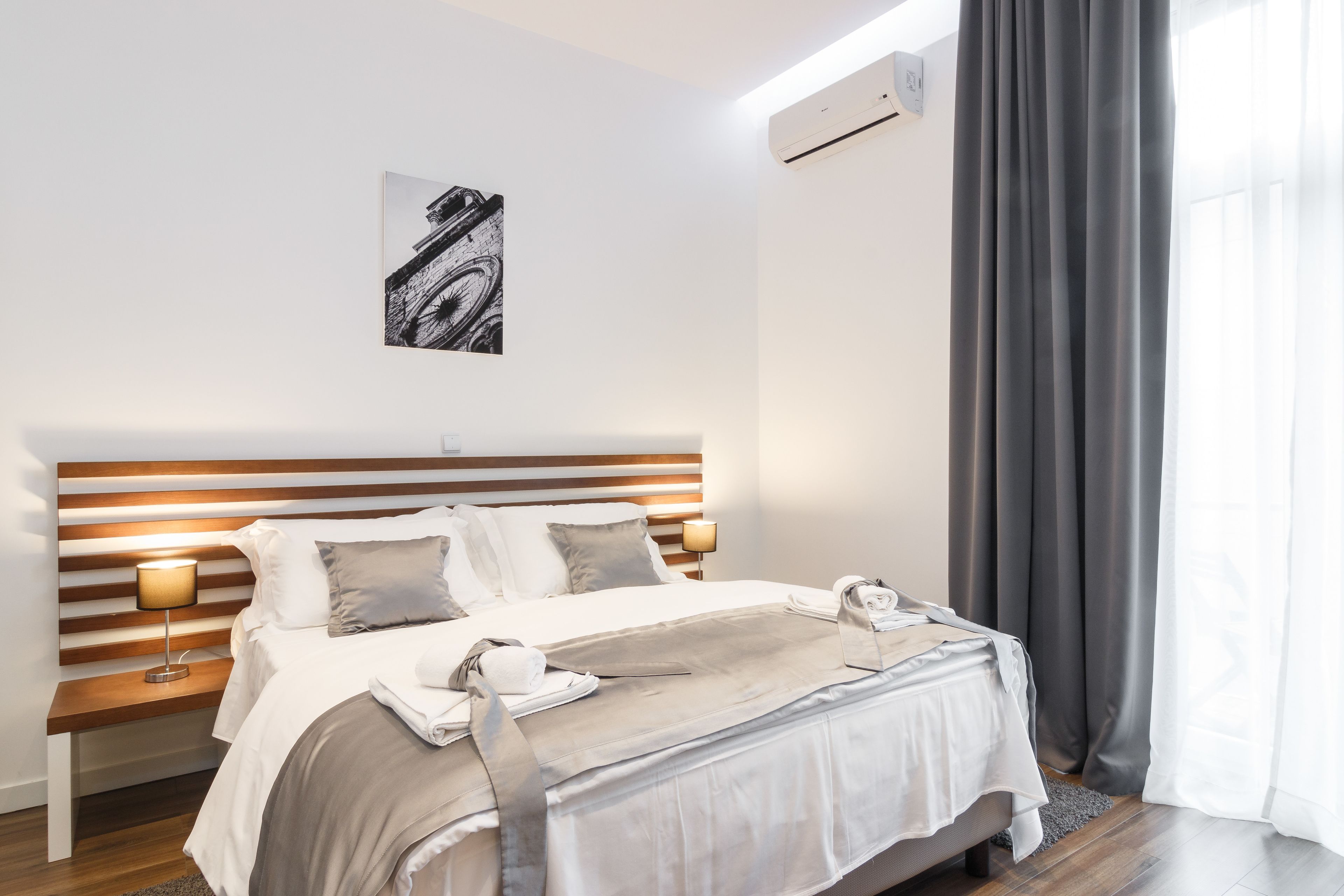 A modern hotel room with a wooden headboard, a bed with white linens, a bedside lamp, a towel and bag on the bed, and a painting on the wall.