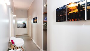 A hallway with white walls, a white chair with a red flower, and a kitchen area in the background.