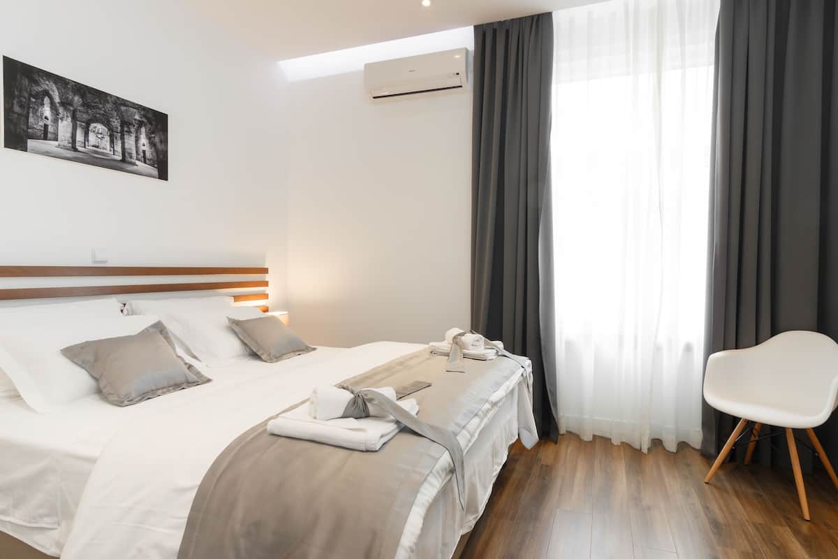 A neatly made bed with a headboard, a bedside table, a chair, and a painting on the wall.