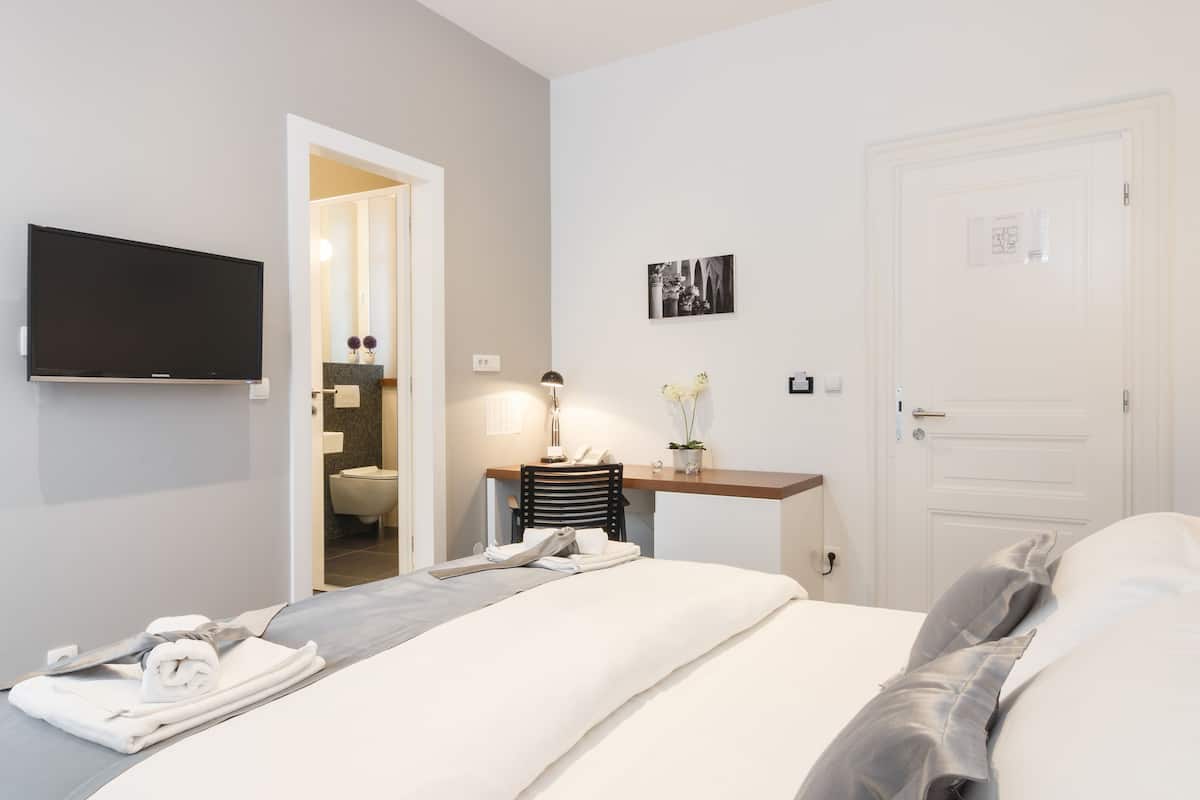 A hotel room with a bed, a desk with a lamp, a TV mounted on the wall, and a door.