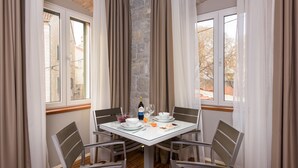 A dining area with a table set for four, including plates, bowls, and glasses. A bottle of wine and a glass are on the table. The room has stone walls and large windows with curtains.