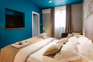 A hotel room with a large bed, a flat-screen TV mounted on the wall, a bedside table with a lamp, and a window with curtains.