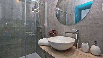 Un baño moderno con ducha de vidrio, un espejo redondo grande y un tocador de madera con lavabo tipo cuenco blanco.