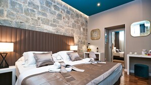 A modern hotel room with a stone wall, a large bed with a headboard, bedside tables with lamps, a desk with a chair, and a mirror on the wall.