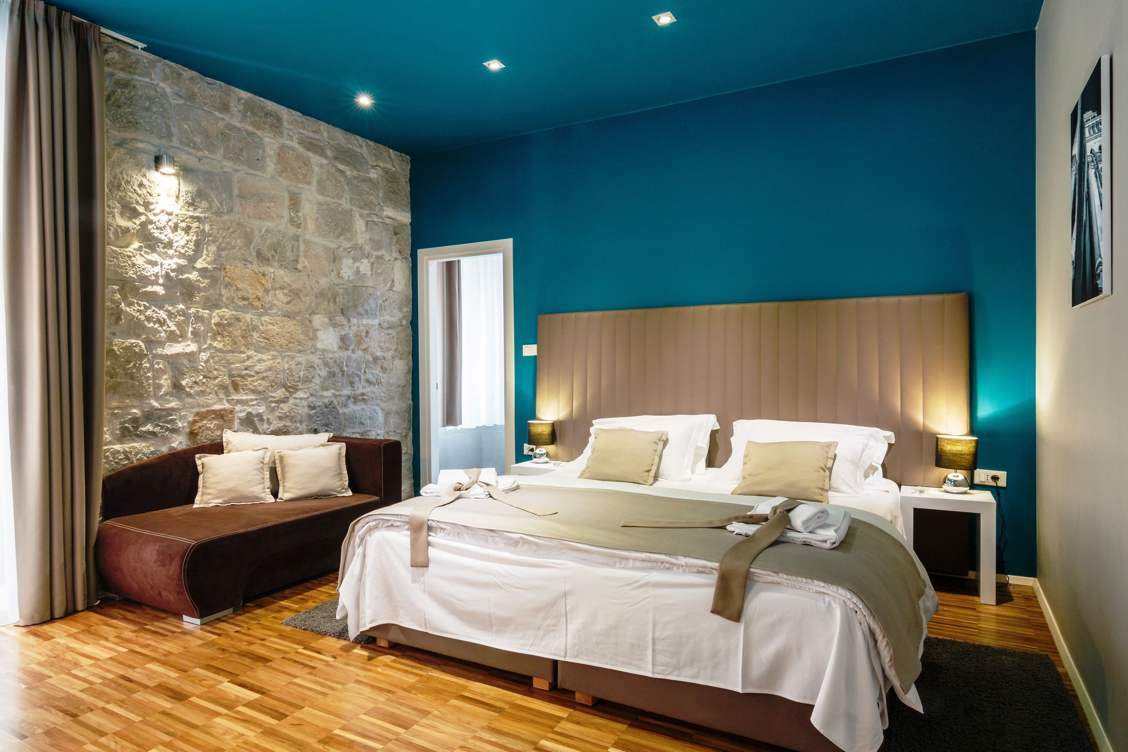 Un dormitorio moderno con pared de piedra decorativa, una cama grande con cabecera de madera, un sofá marrón y una mesita de noche con lámpara.