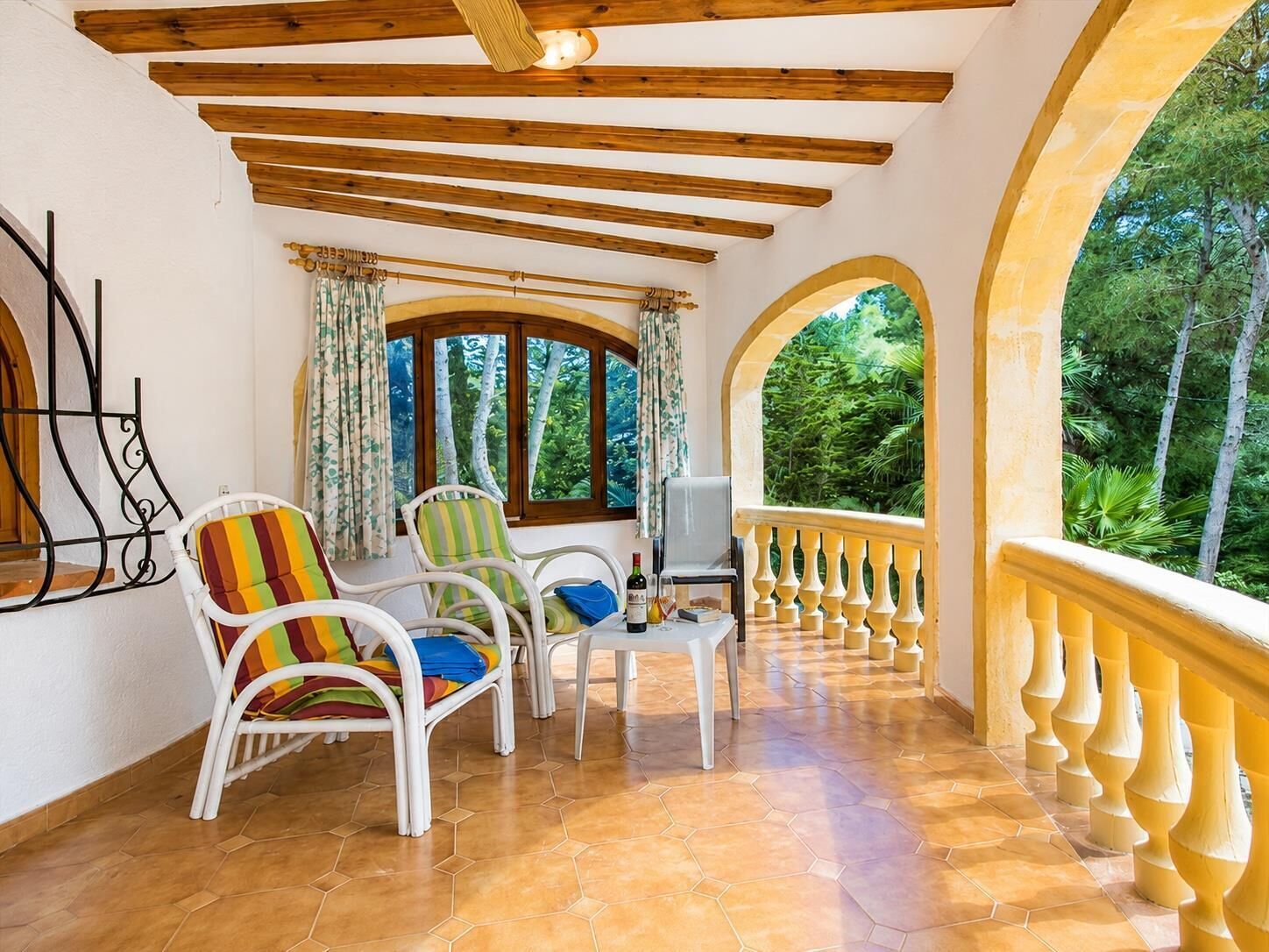 Un patio techado con sillas de mimbre blanco, piso de baldosas y vista a la vegetación a través de un arco.