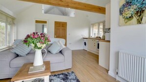 Un salon moderne avec un canapé gris, une table basse blanche et un vase de fleurs. Une poutre en bois est visible au plafond, et on aperçoit une cuisine au fond.