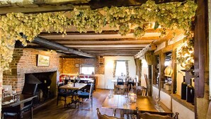 Un coin repas rustique avec des murs en briques apparentes, des poutres en bois et une cheminée en briques. Des tables et des chaises sont disposées pour les repas, et des verres et des bouteilles de vin sont posés sur les tables.