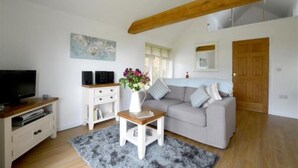 Un salon avec un canapé gris, une petite table blanche ornée d’un vase de fleurs, un téléviseur posé sur un meuble en bois et une poutre en bois au plafond.