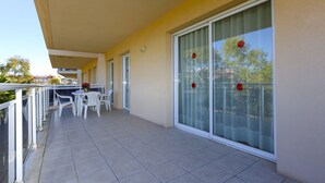 Ein Balkon mit weißen Stühlen und einem Tisch, eine Schiebeglastür und Blick auf Bäume und Gebäude.