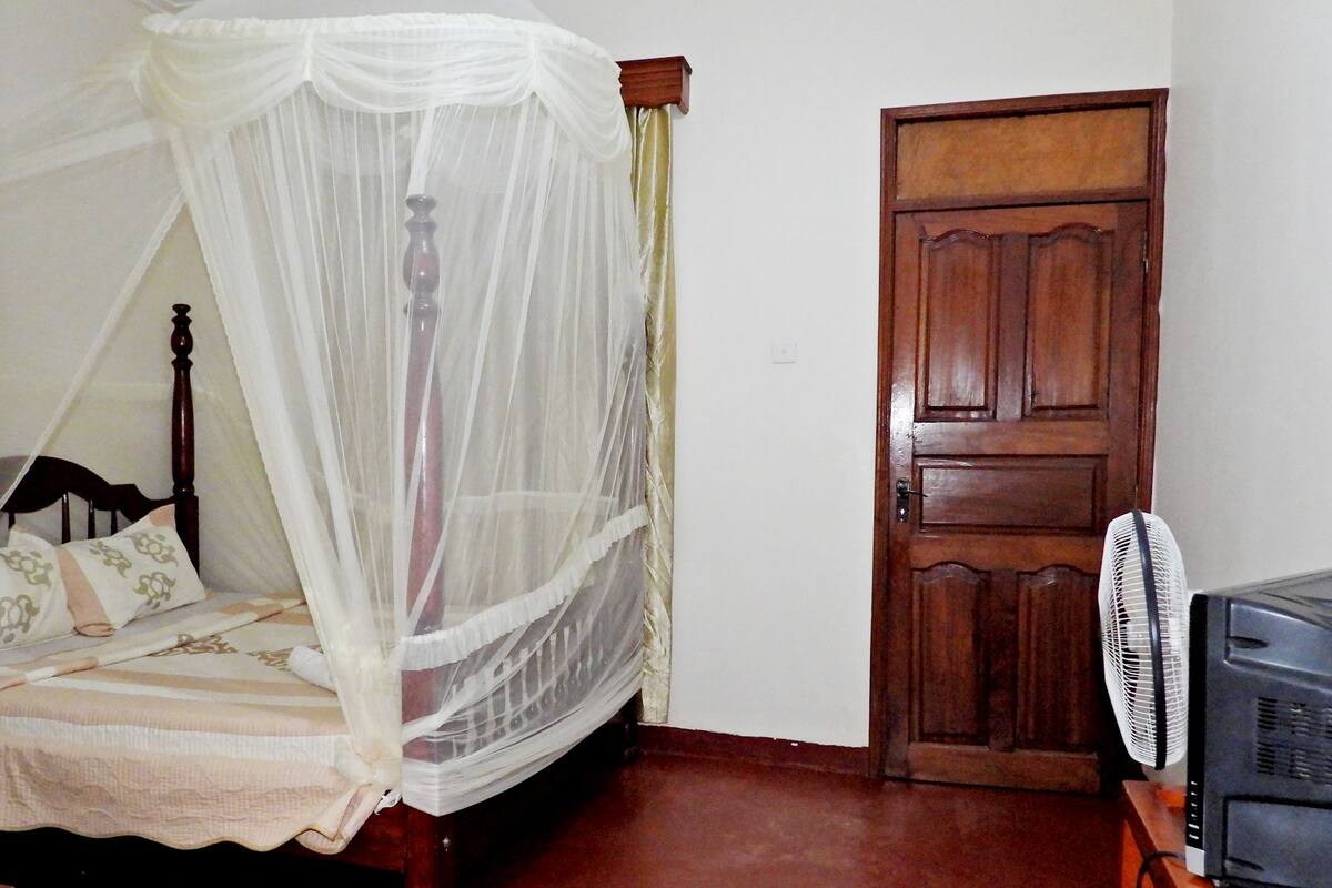A bedroom with a four-poster bed, a wooden door, and a standing fan.