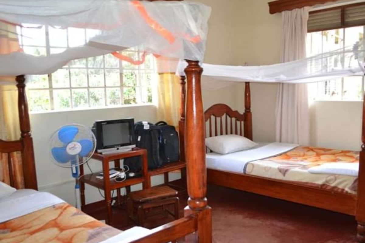 A room with two single beds, a wooden nightstand with a fan, a desk with a computer, and a window with curtains.