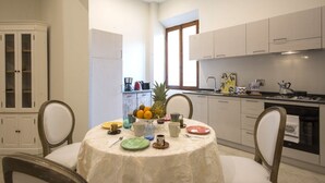 Zona pranzo con tavolo rotondo, sedie, una ciotola di frutta e pensili da cucina sullo sfondo.