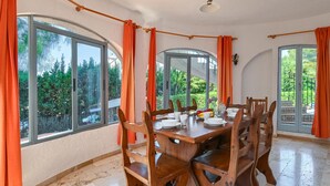 A dining area with a wooden table and chairs, large windows with curtains, and a view of greenery outside. - Escabusso 6 pax - Javea, Costa Blanca (Javea)
