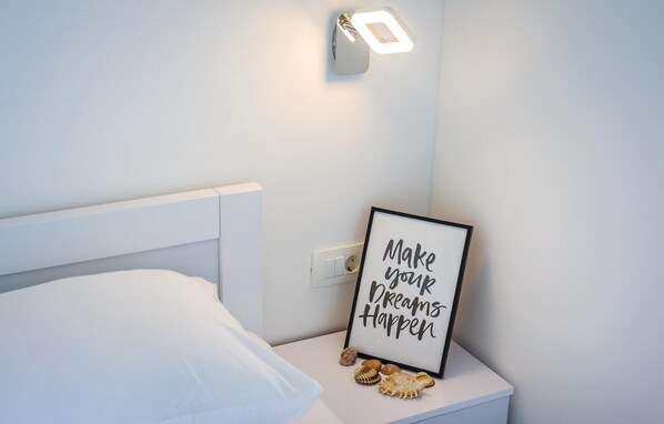 Een net opgemaakt bed met een wit hoofdeinde, een wandlamp en een geïnspireerd citaat in een lijst op een nachtkastje.