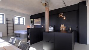 Una cocina moderna con una isla central, armarios negros e iluminación colgante. Hay una escalera apoyada contra la pared y una ventana que proporciona luz natural.