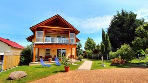 Une maison en bois à deux étages, dotée d’un balcon, d’une allée pavée et d’un jardin bien entretenu avec des plantes en pot et une pelouse.