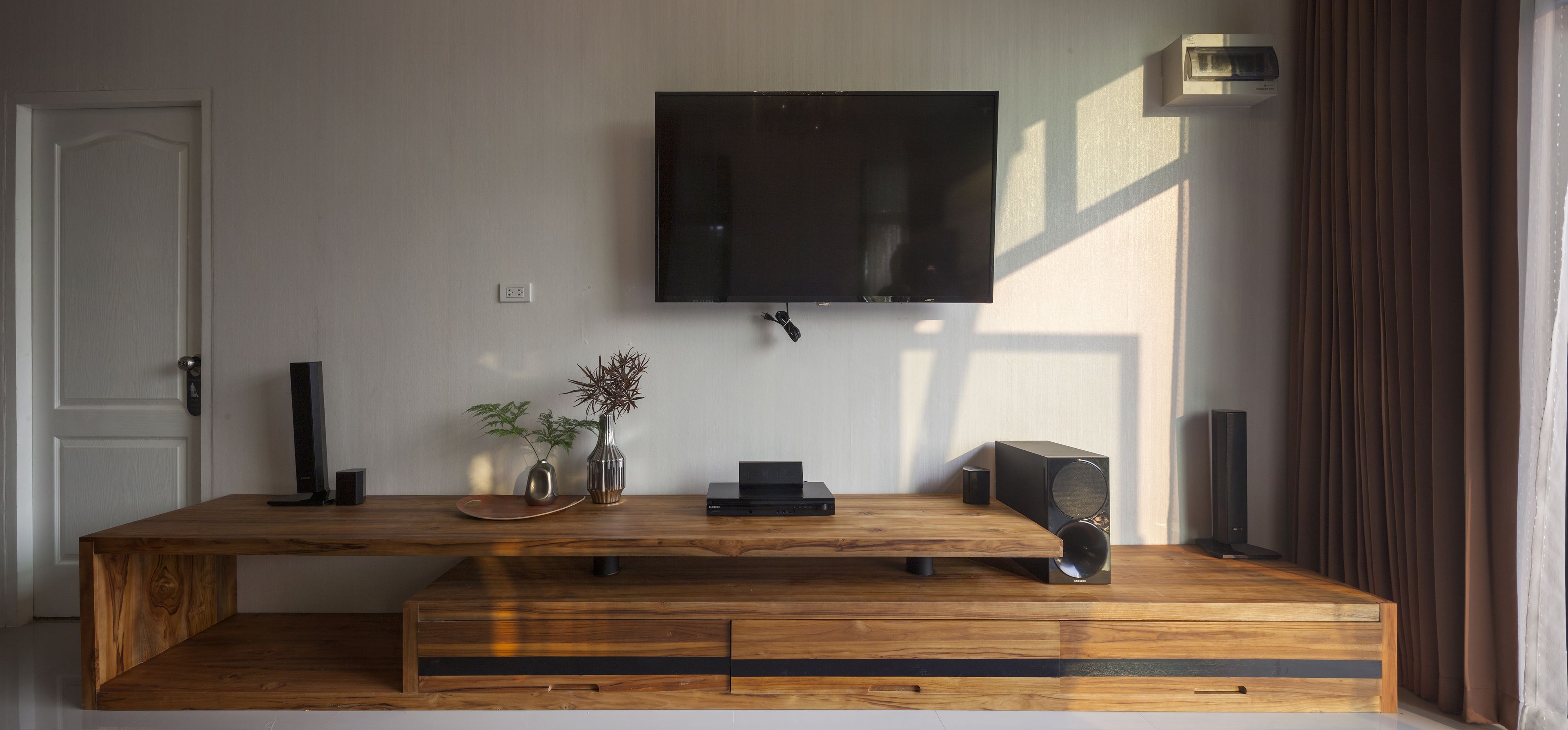 A modern living room with a wooden entertainment center, a flat-screen TV mounted on the wall, and a large window with curtains.