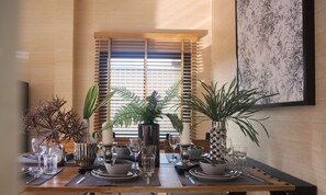 A dining table set with plates, bowls, glasses, and cutlery, flanked by potted plants and a window with blinds.