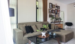 A living room with a sofa, a coffee table, and a plant. There are framed pictures on the wall and a mirror.